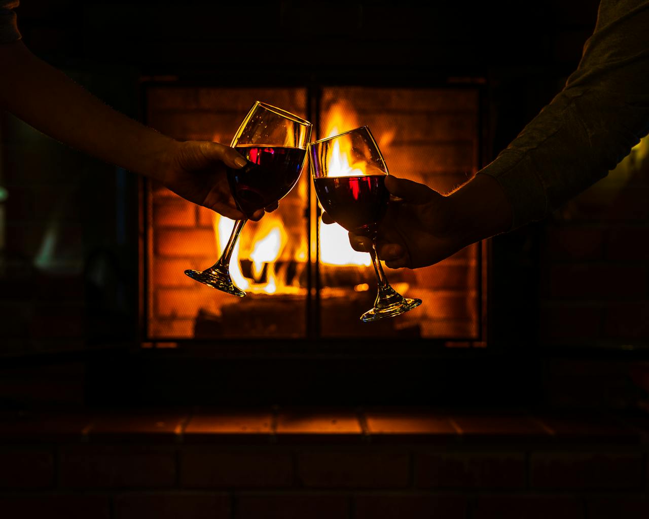 Two individuals toasting with red wine glasses beside a warm fireplace, creating a cozy ambiance.